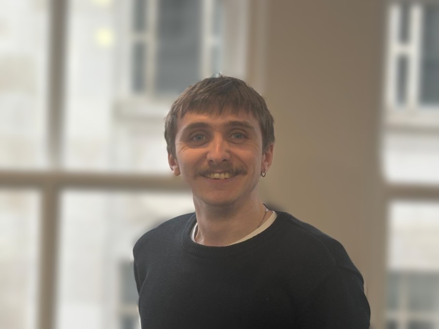 A smiling man with a mustache, wearing a black sweater, stands in an indoor setting with large windows in the background.