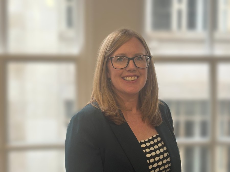 A smiling woman with shoulder-length hair wearing glasses and a black blazer over a polka dot shirt, standing in an indoor setting with large windows in the background.